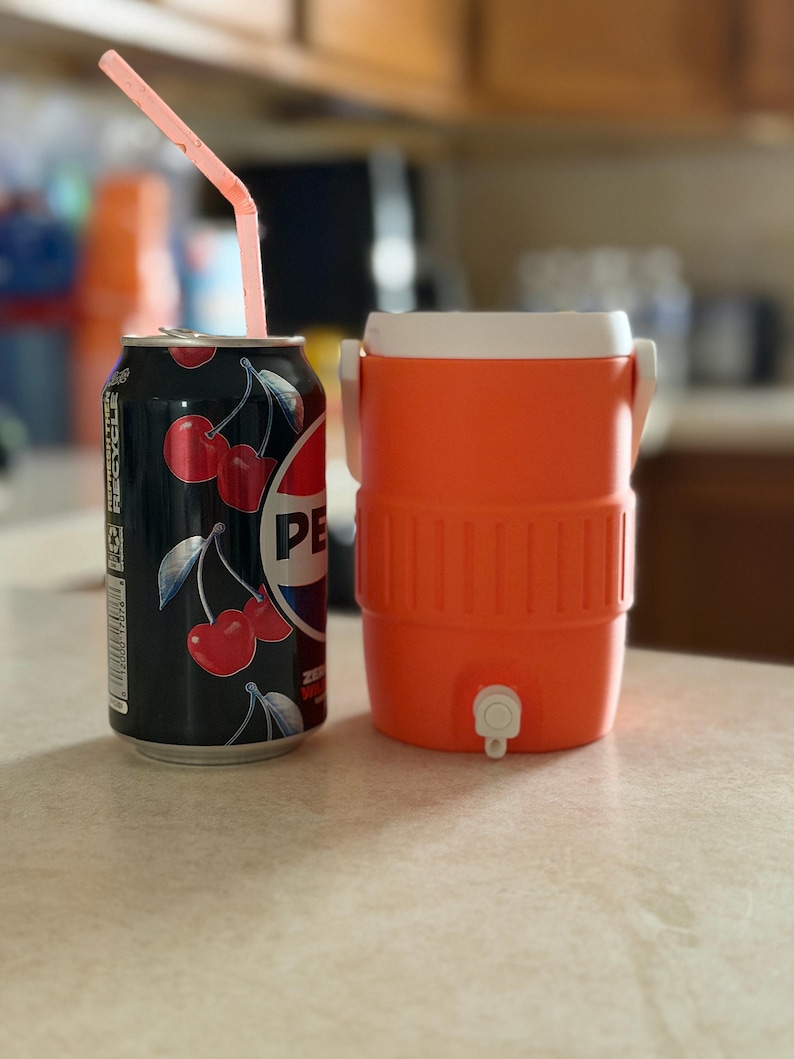 May include: A black cherry-flavored Pepsi can with a pink straw next to an orange and white cooler. The can features a cherry design and the Pepsi logo. The cooler has a spigot and a white lid.
