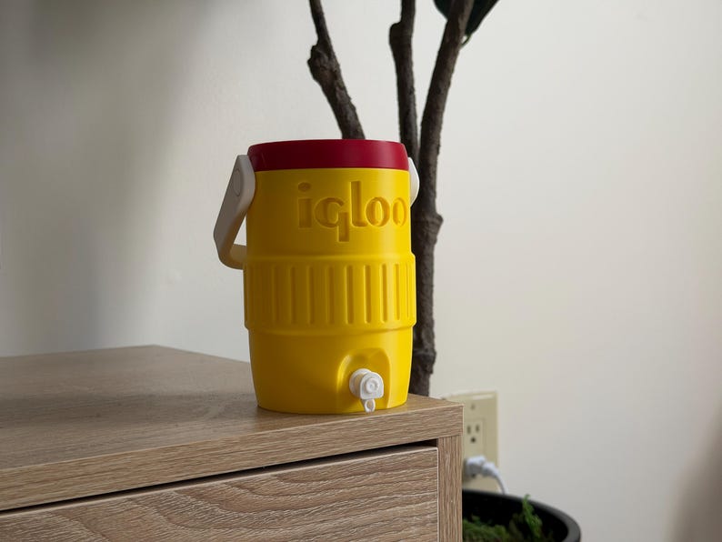 May include: A yellow Igloo water cooler with a red lid and white handle. The cooler has a spigot and the Igloo logo in yellow. The cooler is sitting on a wooden surface.