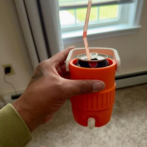 May include: An orange beverage cooler with a white handle holds a soda can with a straw. The cooler has a small white spigot at the bottom. The can is black with red accents.