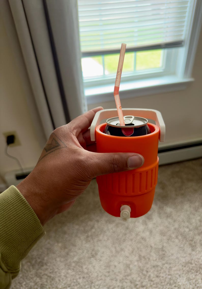 May include: An orange beverage cooler with a white handle holds a soda can with a straw. The cooler has a small white spigot at the bottom. The can is black with red accents.