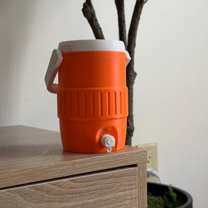 May include: An orange and white water cooler with a spigot. The cooler has a white handle and lid. The cooler is sitting on a wooden surface.