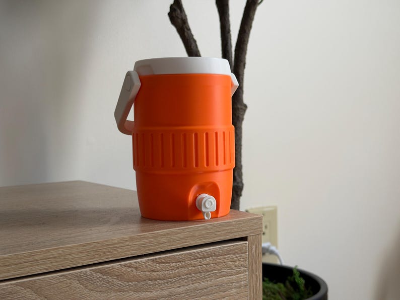 May include: An orange and white water cooler with a spigot. The cooler has a white handle and lid. The cooler is sitting on a wooden surface.