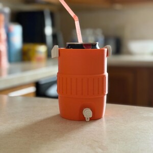 May include: An orange beverage cooler with a white spigot and handles, holding a can with a pink straw. The cooler is cylindrical with a ribbed design around the middle. The straw is bent at an angle.