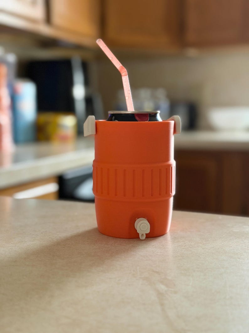 May include: An orange beverage cooler with a white spigot and handles, holding a can with a pink straw. The cooler is cylindrical with a ribbed design around the middle. The straw is bent at an angle.