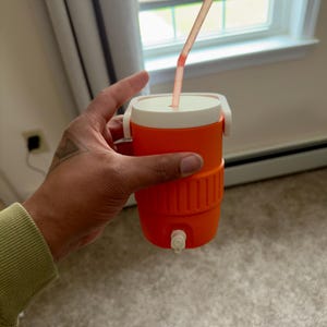 May include: An orange and white insulated beverage cooler with a straw. The cooler has a white lid and handle, and a spigot at the bottom. The straw is pink and extends from the top. Perfect for keeping drinks cold.