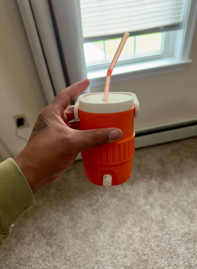 May include: An orange and white insulated beverage cooler with a straw. The cooler has a white lid and handle, and a spigot at the bottom. The straw is pink and extends from the top. Perfect for keeping drinks cold.