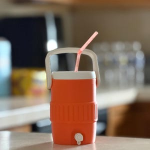 May include: An orange and white cooler with a white handle and a pink straw. The cooler has a spigot at the bottom and is sitting on a light-colored surface. The cooler is designed to keep drinks cold.