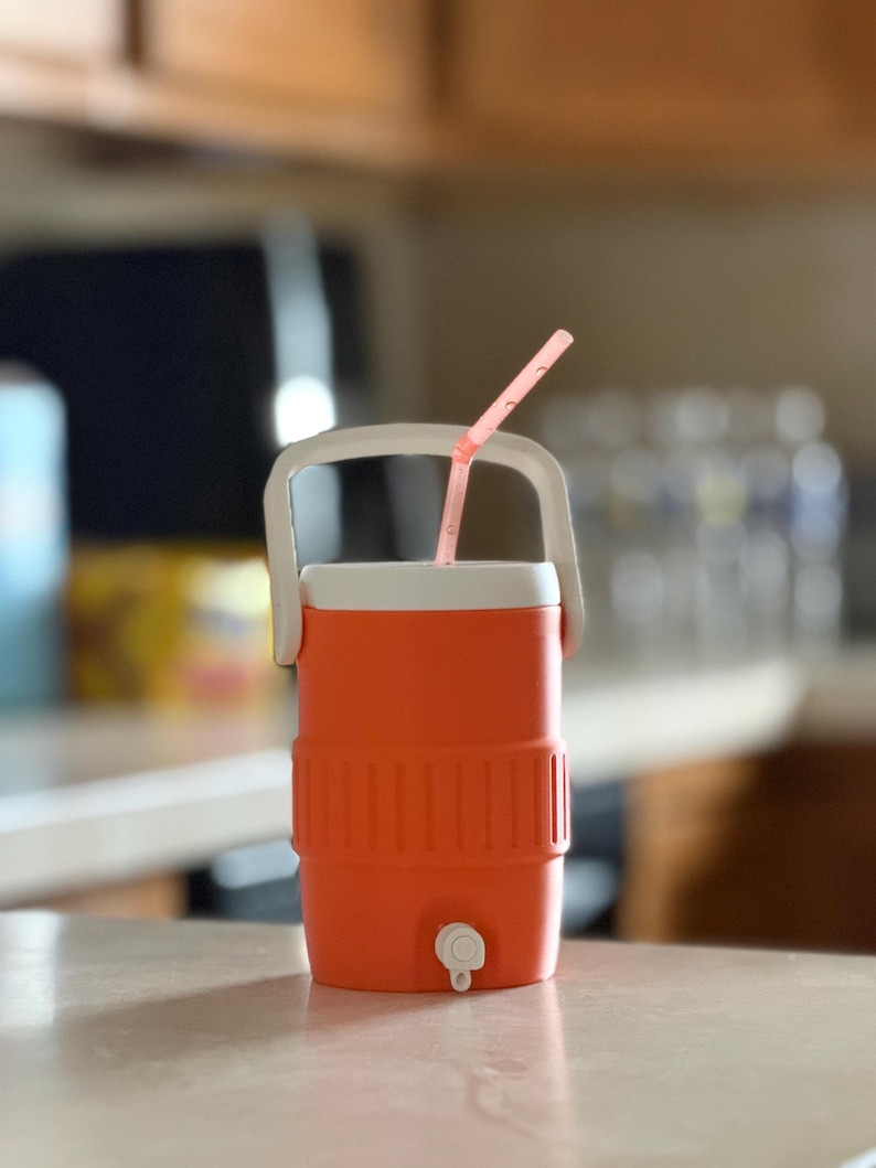 May include: An orange and white cooler with a white handle and a pink straw. The cooler has a spigot at the bottom and is sitting on a light-colored surface. The cooler is designed to keep drinks cold.