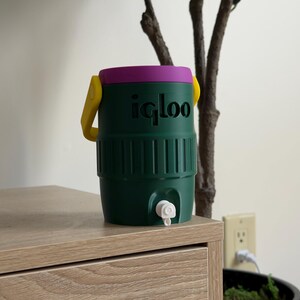 May include: A green Igloo water cooler with a purple lid and yellow handle. The cooler has a white spigot and is sitting on a wooden surface. The Igloo logo is printed in black on the front of the cooler.