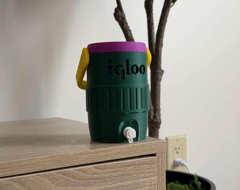 May include: A green Igloo water cooler with a purple lid and yellow handle. The cooler has a white spigot and is sitting on a wooden surface. The Igloo logo is printed in black on the front of the cooler.