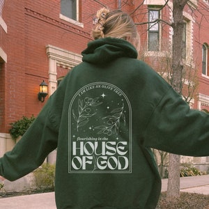 Puede incluir: Una sudadera con capucha verde oscuro con un dise&ntilde;o gr&aacute;fico blanco que presenta un olivo y el texto "Soy como un olivo que florece en la casa de Dios".