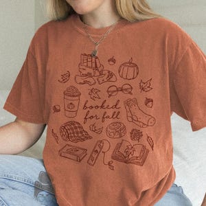 May include: A rust-coloured short-sleeve t-shirt featuring a dark brown line art graphic of autumn-themed items including books, a coffee cup, glasses, a sock, and a cinnamon roll with the text "booked for fall" printed in the centre.