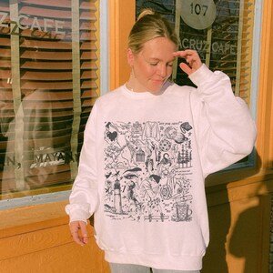 Könnte beinhalten: Weißer Pullover mit einem schwarz-weißen Grafikdruck verschiedener Illustrationen, darunter ein Leuchtturm, ein Vogel, ein Herz und ein Stern. Der Text "Are you happy?" ist auf dem Pullover gedruckt.