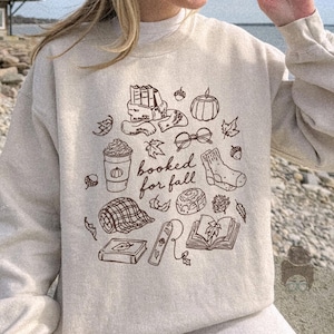 Puede incluir: Una sudadera beige de cuello redondo con un gráfico de líneas marrones en la parte delantera. El diseño incluye una pila de libros, una taza de café, una calabaza, gafas, un calcetín, una manta a cuadros y el texto "booked for fall" en cursiva.