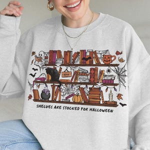 Op de afbeelding: Een asgrijze sweater met ronde hals en een grafische print van drie houten planken vol boeken, een zwarte kat, een geest, een pompoen, een ketel en spinnenwebben. De tekst onder de planken luidt: "SHELVES ARE STOCKED FOR HALLOWEEN."