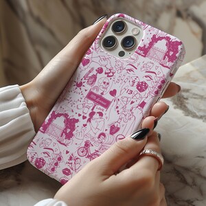 May include: A white iPhone case with a pink and white design featuring hearts, skulls, dice, and other symbols. The case has a camera lens cutout and a pink border.
