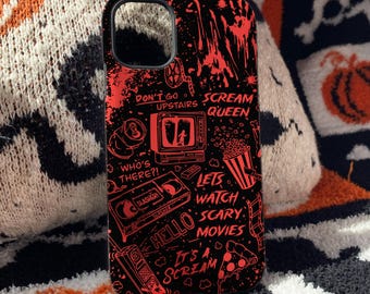 Puede incluir: Una funda de teléfono negra con ilustraciones gráficas rojas de temas de películas de terror, incluyendo un televisor, una cinta VHS, un teléfono, palomitas y pizza, con el texto "Don't go upstairs," "Scream Queen," "Who's there?," "Hello," y "Let's watch scary movies."