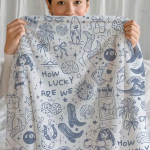 May include: A white throw blanket with a blue and white pattern. The pattern includes palm trees, stars, cherries, dice, horseshoes, a disco ball, a surfboard, and the text "How lucky are we".