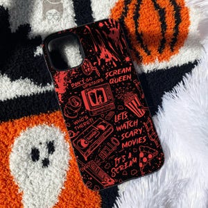 May include: A black phone case featuring a red graphic print of horror movie motifs including a television, a VHS tape, a popcorn bucket, and a slice of pizza. The text reads: Don't go upstairs, Scream Queen, Who's there?, Lets watch scary movies, Hello, It's a scream.