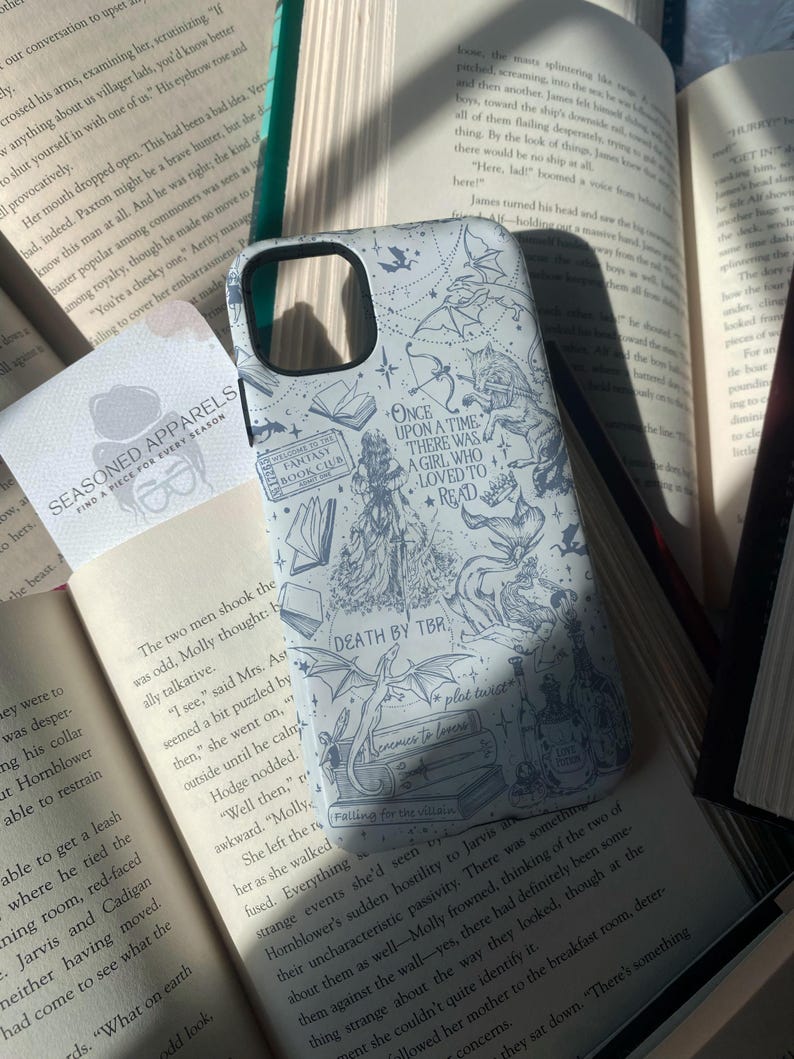 May include: A light-colored phone case with a fantasy theme, featuring illustrations of dragons, books, and the text "Once Upon a Time There Was a Girl Who Loved to Read." The case is surrounded by open books.