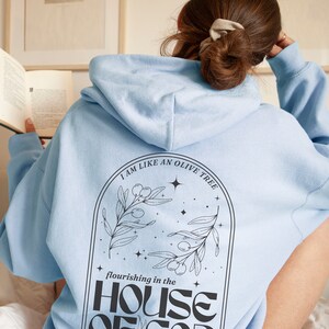 Puede incluir: Sudadera con capucha azul claro con el texto "Soy como un olivo que florece en la casa de Dios" y una ilustraci&oacute;n en blanco y negro de un olivo con estrellas.