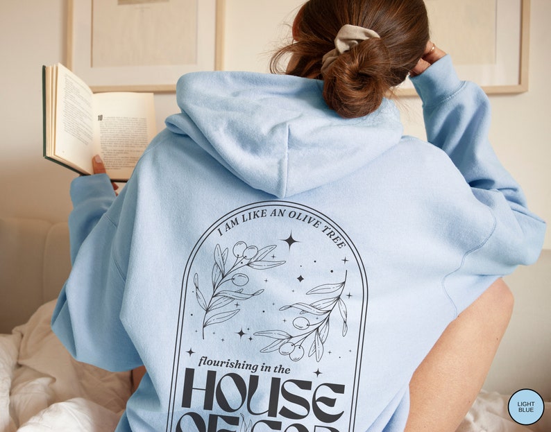 Puede incluir: Sudadera con capucha azul claro con el texto "Soy como un olivo que florece en la casa de Dios" y una ilustraci&oacute;n en blanco y negro de un olivo con estrellas.