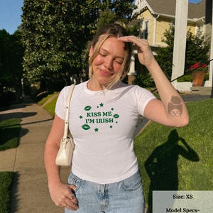 May include: White t-shirt with the text "KISS ME I'M IRISH" in green, along with green shamrock and star graphics. The shirt is paired with light blue jeans. A white purse hangs from the shoulder.