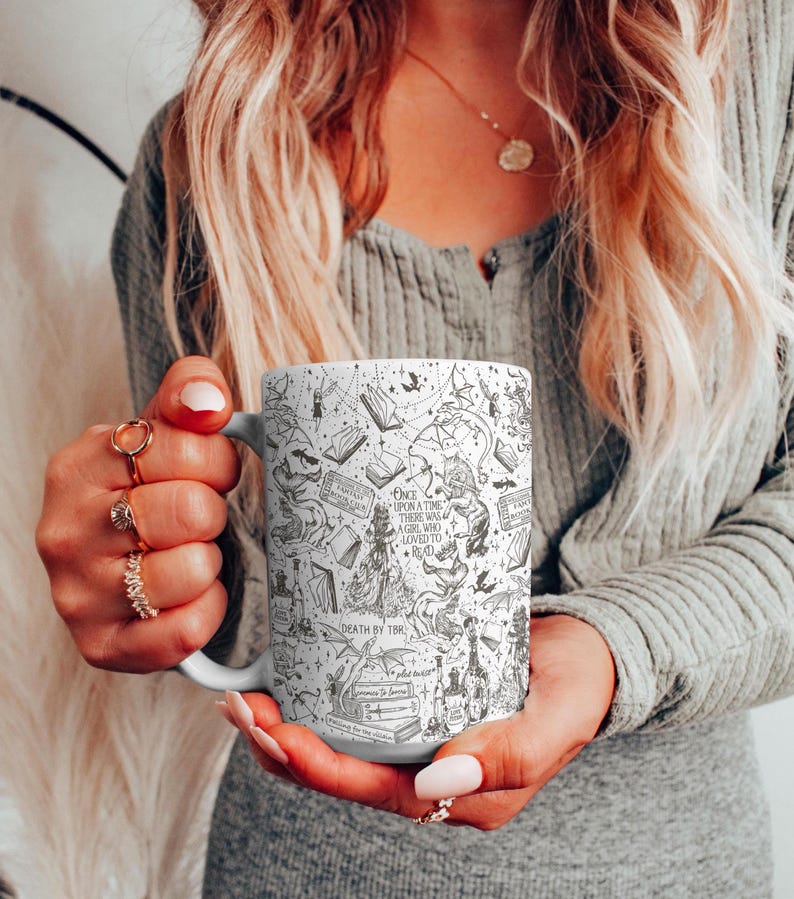 K&ouml;nnte beinhalten: Eine wei&szlig;e Keramiktasse mit einer Schwarzwei&szlig;illustration eines M&auml;dchens, das ein Buch liest, umgeben von magischen Elementen wie Drachen, Eulen und Tr&auml;nken. Der Text auf der Tasse lautet "Once upon a time there was a girl who loved to read. Death by TBR."