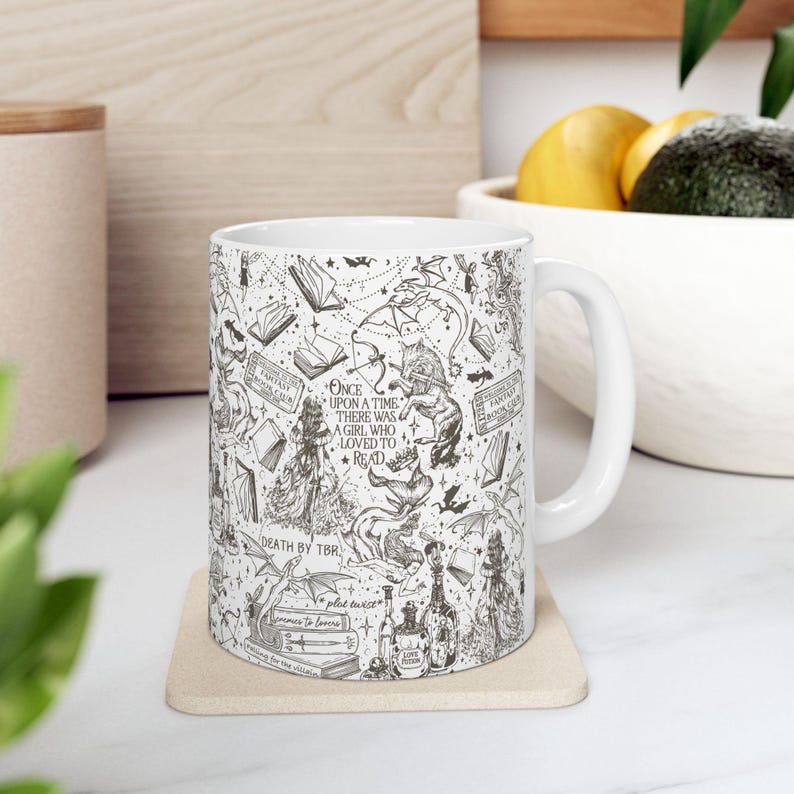 K&ouml;nnte beinhalten: Wei&szlig;e Keramiktasse mit einer schwarz-wei&szlig;en Illustration eines Buch-Designs. Das Design beinhaltet einen Drachen, ein Buch und den Text "Es war einmal ein M&auml;dchen, das es liebte zu lesen".