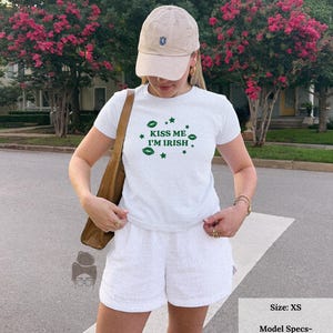 May include: White t-shirt with the text "KISS ME I'M IRISH" in green, paired with white shorts and a beige baseball cap. The image shows a casual outfit, perfect for St. Patrick's Day or any Irish-themed event.