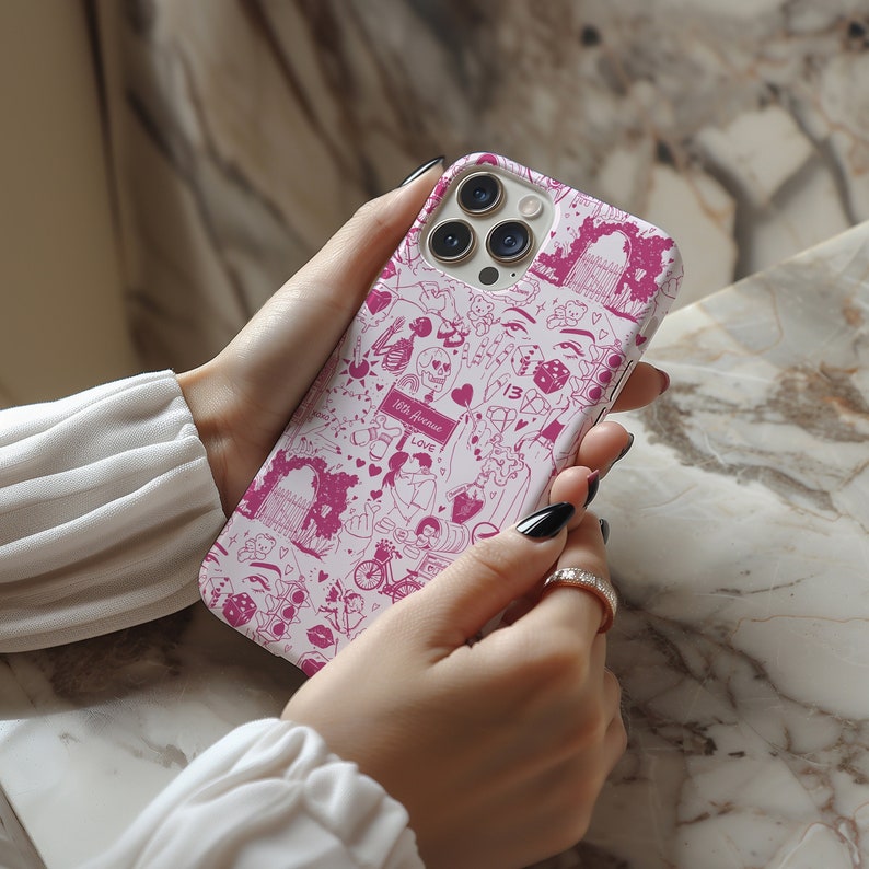 May include: A white iPhone case with a pink and white design featuring hearts, skulls, dice, and other symbols. The case has a camera lens cutout and a pink border.