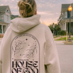 Puede incluir: Una sudadera con capucha beige con un dise&ntilde;o gr&aacute;fico negro en la espalda. El gr&aacute;fico presenta un olivo con el texto "I am like an olive tree flourishing in the HOUSE OF GOD".