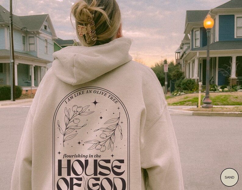 Puede incluir: Una sudadera con capucha beige con un dise&ntilde;o gr&aacute;fico negro en la espalda. El gr&aacute;fico presenta un olivo con el texto "I am like an olive tree flourishing in the HOUSE OF GOD".
