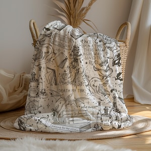 May include: A cream-colored throw blanket with a black and white print of books, potions, and dragons. The text on the blanket reads "Welcome to the Fantasy Book Club", "Once upon a time, there was a girl who loved to read", "Death by TBR", "*plot twist*", "Enemies to lovers", "Falling for the villain", and "Love Potion".