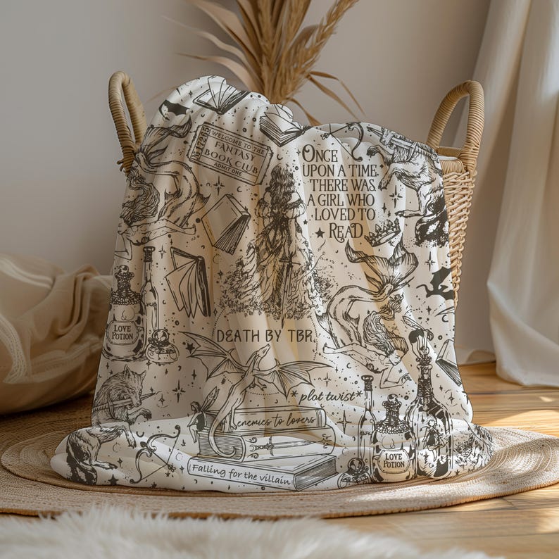 May include: A cream-colored throw blanket with a black and white print of books, potions, and dragons. The text on the blanket reads "Welcome to the Fantasy Book Club", "Once upon a time, there was a girl who loved to read", "Death by TBR", "*plot twist*", "Enemies to lovers", "Falling for the villain", and "Love Potion".