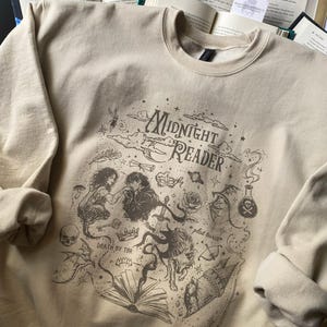 Op de afbeelding: Een beige sweater met ronde hals en een donkerbruine grafische print met de tekst "Midnight Reader" en "Death by TBR". Het ontwerp bevat illustraties van een koppel, een wolf, een zwaard, een kasteel, een schedel en een opengeslagen boek.