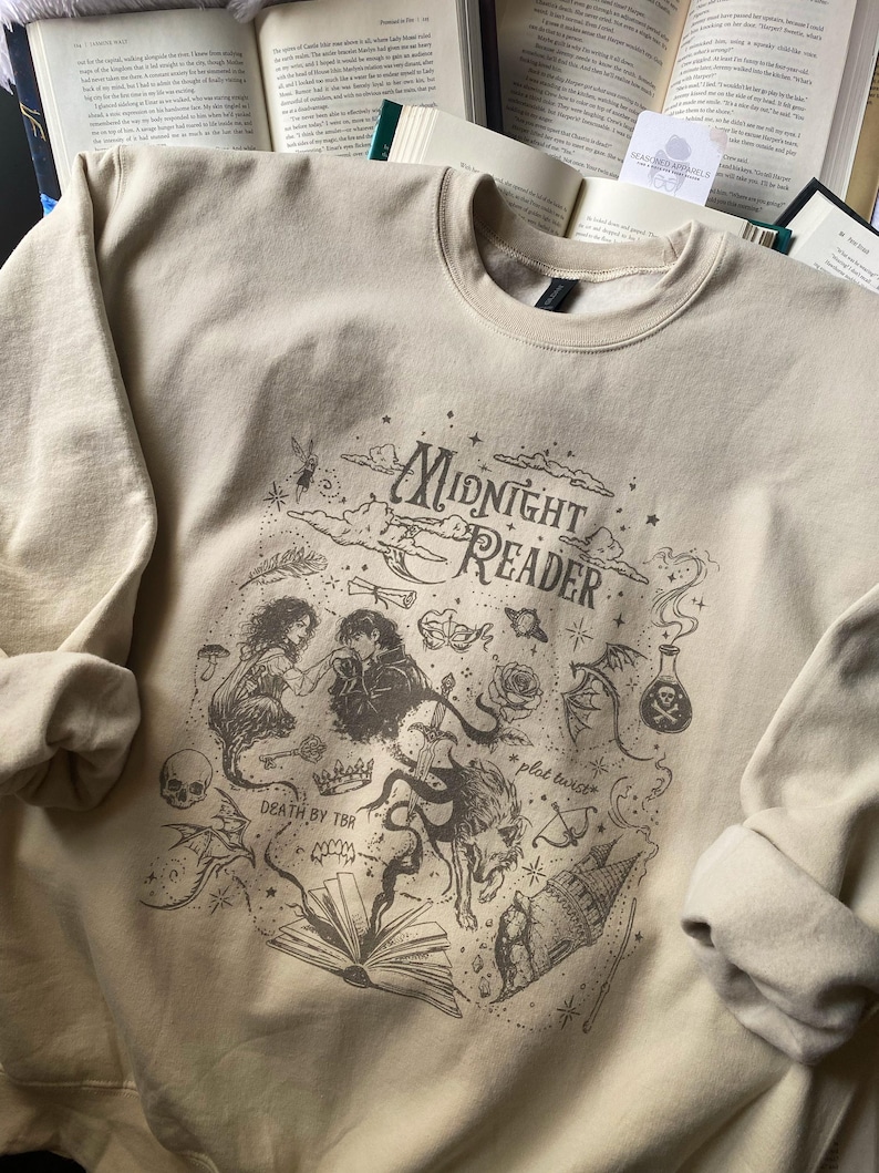 Op de afbeelding: Een beige sweater met ronde hals en een donkerbruine grafische print met de tekst "Midnight Reader" en "Death by TBR". Het ontwerp bevat illustraties van een koppel, een wolf, een zwaard, een kasteel, een schedel en een opengeslagen boek.