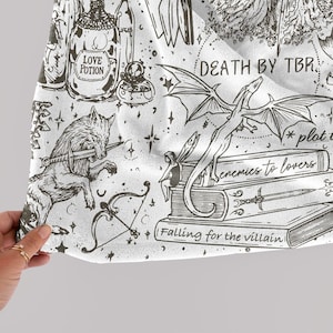 May include: A white fabric with a black and white illustration of a dragon, a wolf, a fairy, a bow and arrow, and a bottle of love potion. The fabric has the text "Death by TBR", "*plot twist*" and "Enemies to lovers" on it.