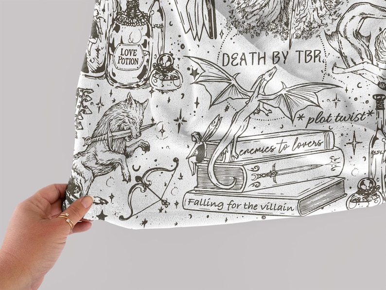 May include: A white fabric with a black and white illustration of a dragon, a wolf, a fairy, a bow and arrow, and a bottle of love potion. The fabric has the text "Death by TBR", "*plot twist*" and "Enemies to lovers" on it.