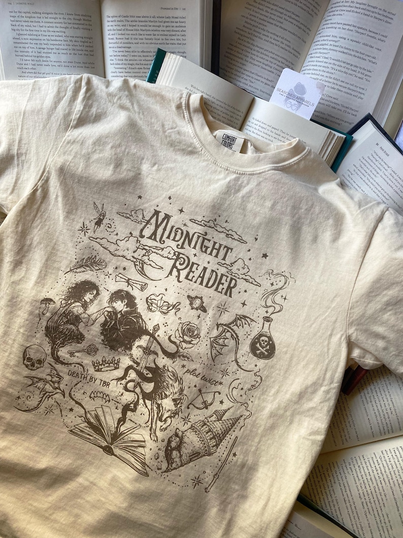 Puede incluir: Una camiseta de manga corta color crema con un estampado gr&aacute;fico marr&oacute;n oscuro con el texto Midnight Reader, Death by TBR y plot twist. El dise&ntilde;o incluye ilustraciones de una pareja, una calavera, una corona, un libro, un castillo y frascos de pociones.