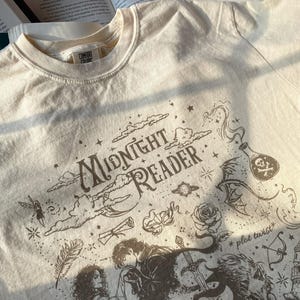 Puede incluir: Una camiseta de color crema con un estampado gr&aacute;fico marr&oacute;n oscuro que incluye el texto "Midnight Reader" y "plot twist". El dise&ntilde;o presenta ilustraciones de una pareja, una espada, una rosa, una botella con una calavera, una corona y estrellas.