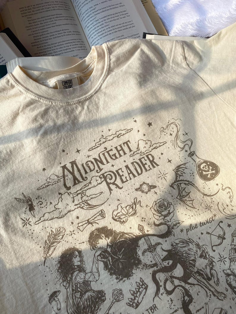 Puede incluir: Una camiseta de color crema con un estampado gr&aacute;fico marr&oacute;n oscuro que incluye el texto "Midnight Reader" y "plot twist". El dise&ntilde;o presenta ilustraciones de una pareja, una espada, una rosa, una botella con una calavera, una corona y estrellas.