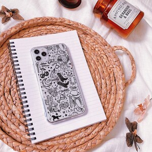Puede incluir: Una funda transparente para tel&eacute;fono con un dise&ntilde;o de vaquero en blanco y negro, con botas, un sombrero y las palabras "Yee Haw", "Howdy" y "Cowboy Tears". La funda descansa sobre un cuaderno espiralado sobre una bandeja tejida, con una vela y un peque&ntilde;o recipiente de vidrio.