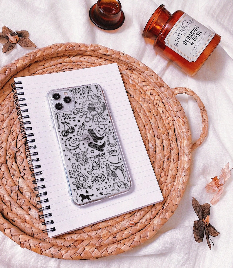 Puede incluir: Una funda transparente para tel&eacute;fono con un dise&ntilde;o de vaquero en blanco y negro, con botas, un sombrero y las palabras "Yee Haw", "Howdy" y "Cowboy Tears". La funda descansa sobre un cuaderno espiralado sobre una bandeja tejida, con una vela y un peque&ntilde;o recipiente de vidrio.