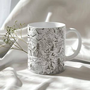 K&ouml;nnte beinhalten: Wei&szlig;e Keramiktasse mit einer Schwarzwei&szlig;illustration von B&uuml;chern, Drachen und anderen Fantasy-Elementen. Der Text auf der Tasse lautet "Es war einmal ein M&auml;dchen, das es liebte zu lesen" und "Fantasy Book Club".