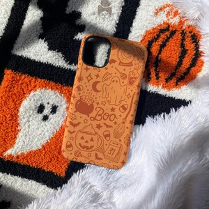 Puede incluir: Funda de tel&eacute;fono naranja con dise&ntilde;o de Halloween. Presenta ilustraciones de un fantasma, una calabaza, una escoba de bruja y la palabra "Boo". El fondo es blanco, negro y naranja.
