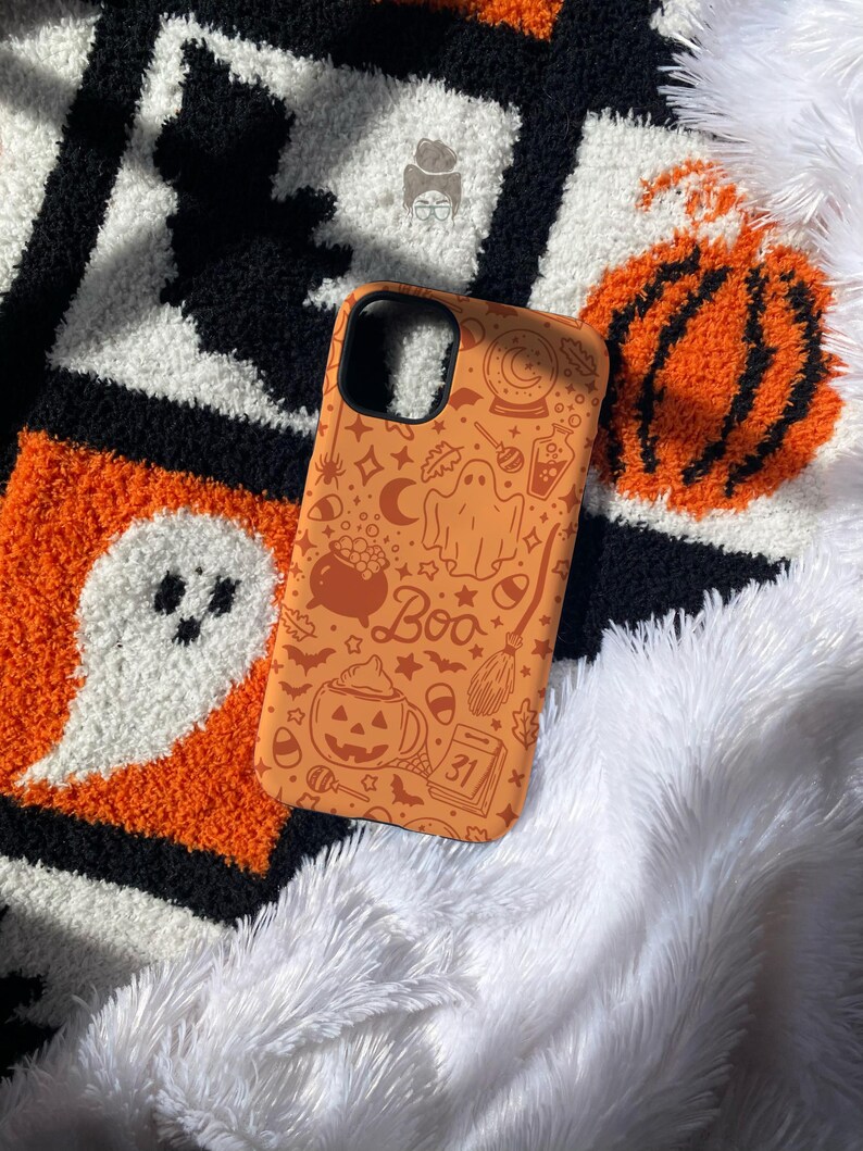 Puede incluir: Funda de tel&eacute;fono naranja con dise&ntilde;o de Halloween. Presenta ilustraciones de un fantasma, una calabaza, una escoba de bruja y la palabra "Boo". El fondo es blanco, negro y naranja.