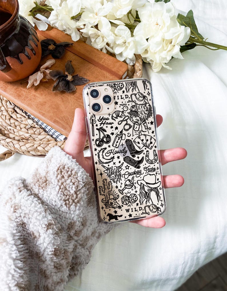 Puede incluir: Funda transparente para tel&eacute;fono con un dise&ntilde;o de tem&aacute;tica vaquera en blanco y negro. La funda presenta ilustraciones de botas de vaquero, un sombrero, un cactus y las palabras "Yee Haw", "Howdy" y "Wild".