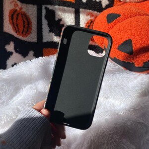 Puede incluir: Funda de tel&eacute;fono negra con acabado mate. La funda se sostiene contra una superficie blanca y esponjosa. El fondo incluye una manta con tem&aacute;tica de Halloween con calabazas, calaveras y fantasmas.