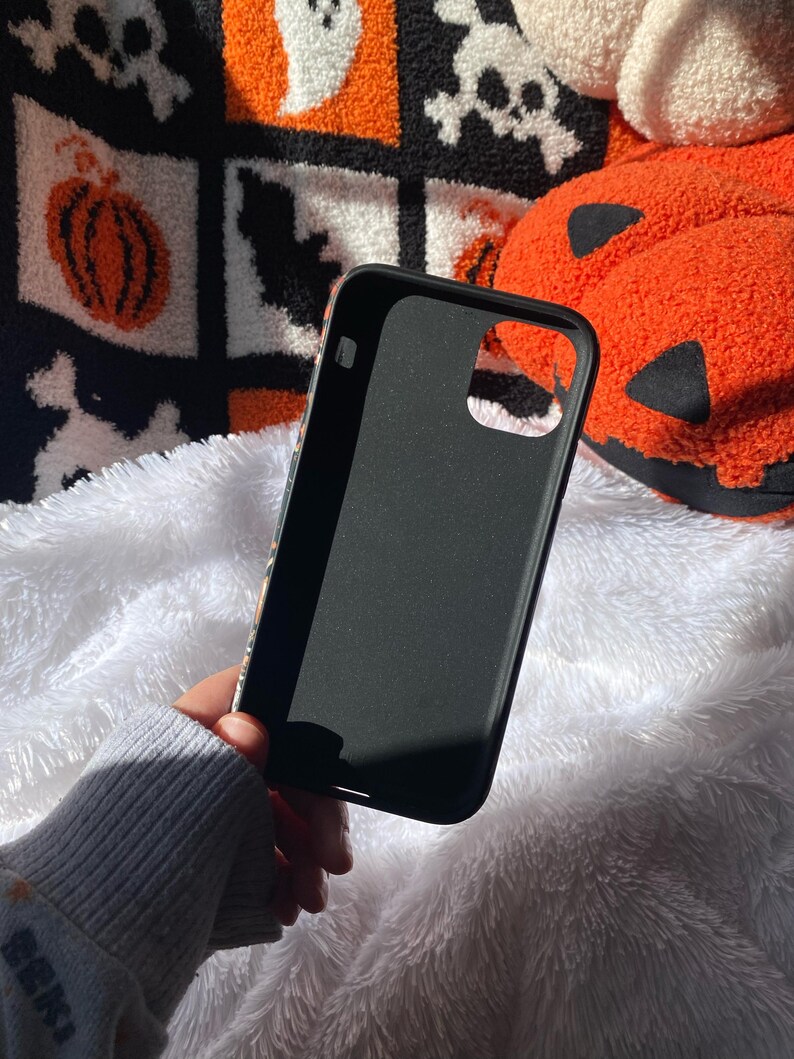 Puede incluir: Funda de tel&eacute;fono negra con acabado mate. La funda se sostiene contra una superficie blanca y esponjosa. El fondo incluye una manta con tem&aacute;tica de Halloween con calabazas, calaveras y fantasmas.